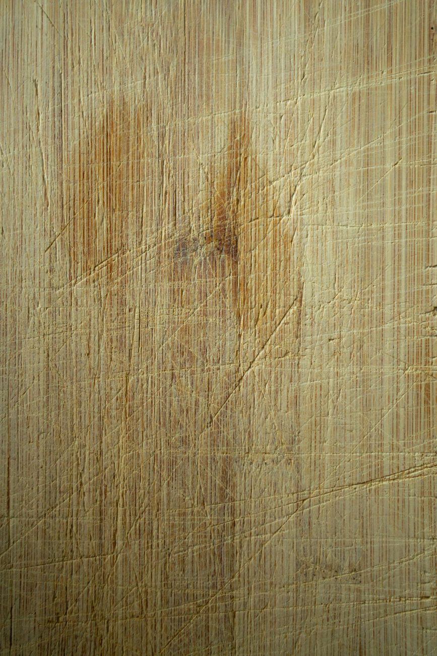 A close-up of a wooden cutting board with visible knife marks and vertical grain — the surface a good board earns over years of daily use.