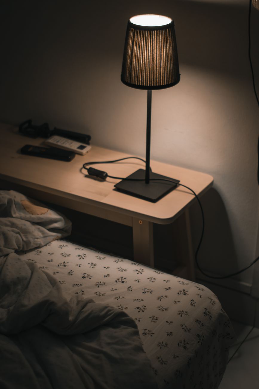 A single bedside lamp lit in an otherwise dark bedroom — phase three of the protocol, where the room reads at roughly 150 lumens and 2200K.