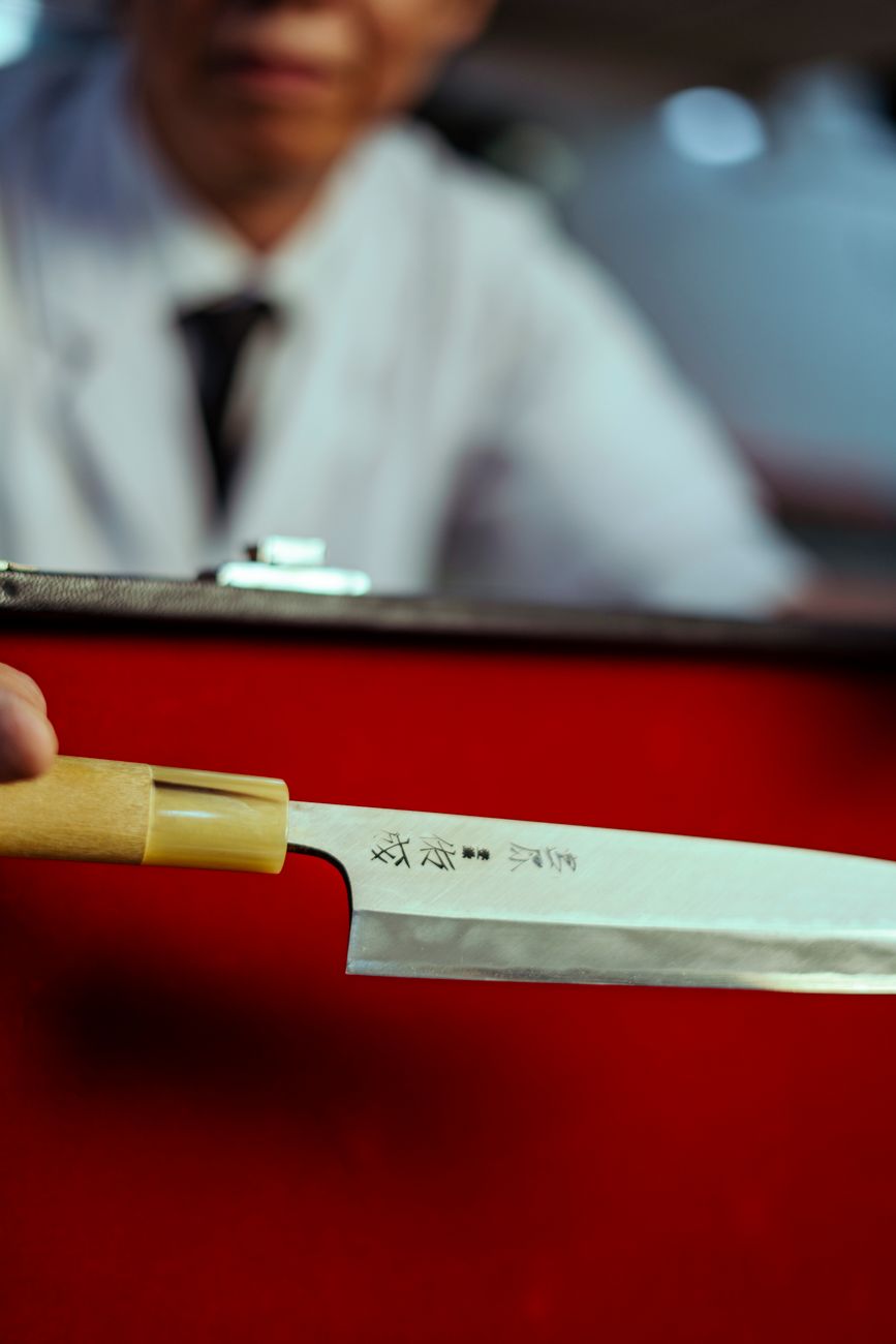 A chef holds a Japanese chef's knife with a wooden handle, the flat of the blade catching light — the kind of craftsmanship the heirloom tier is paying for.