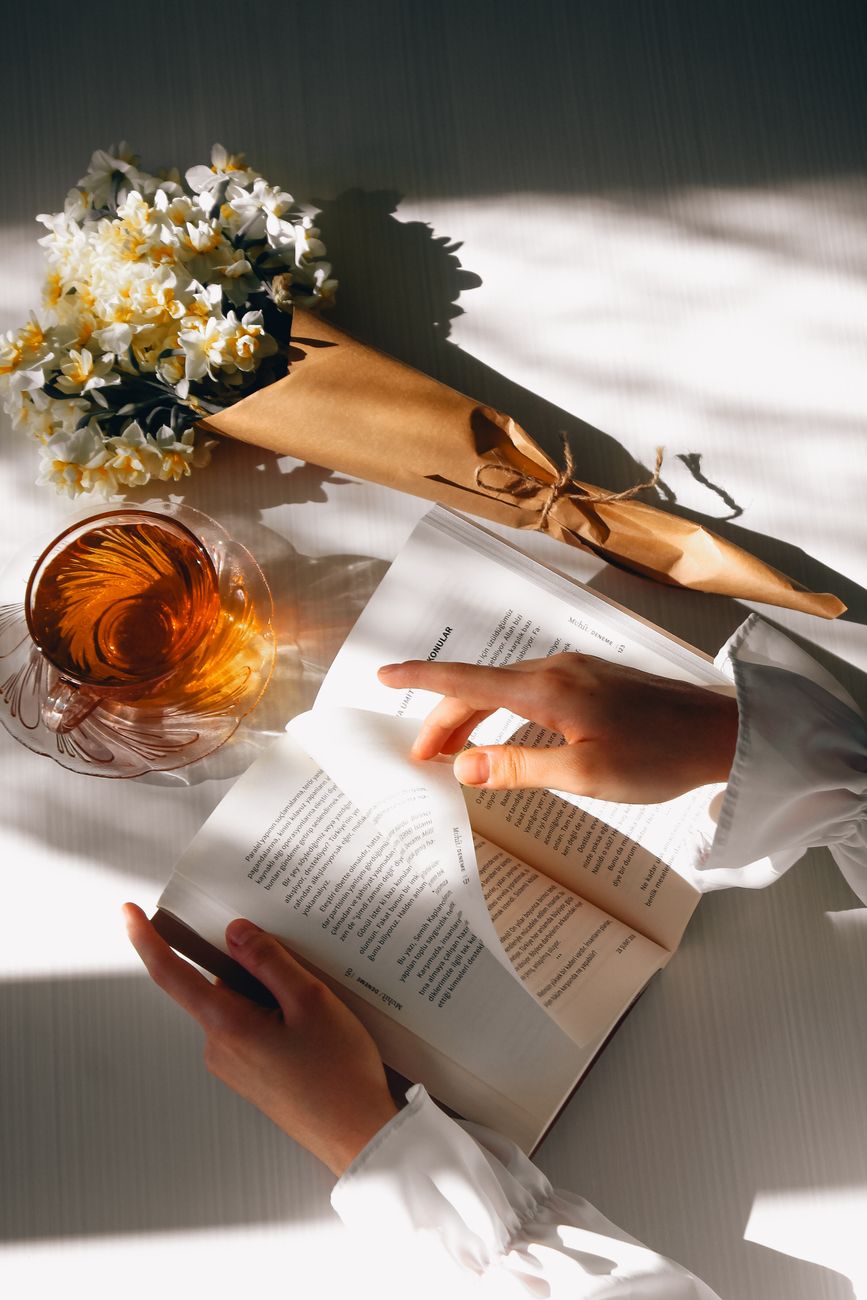 Hands holding a book among fresh flowers and a cup of tea — the editorial position of this publication in a single frame.